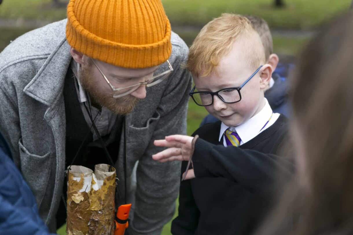 Eco Drama - Out to Play. Boy holds a worm in his hand while a man dressed up as an adventurer with binoculars looks on.