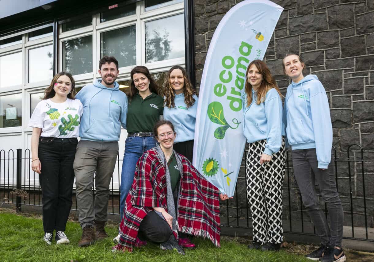 A group of people wearing Eco Drama branding smiling and huddling together for a photo with white banner with Eco Drama written in green text.