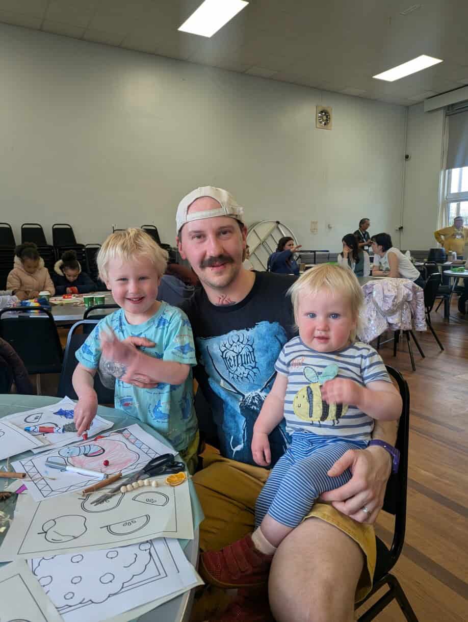 A man sits with his two children on his lap. They are colouring in as a family at a harvest festival event